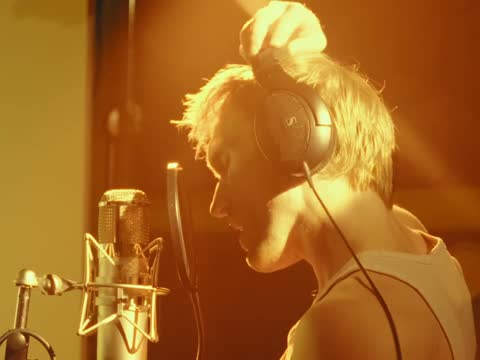 RAW 002, frame 08: the singer in headphones, lit amber, in profile at the microphone