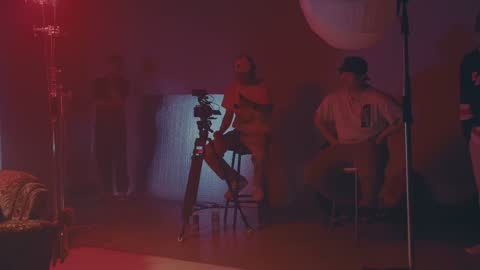 RUINAS BTS, frame 04: two crew members on stools behind a camera on a tripod