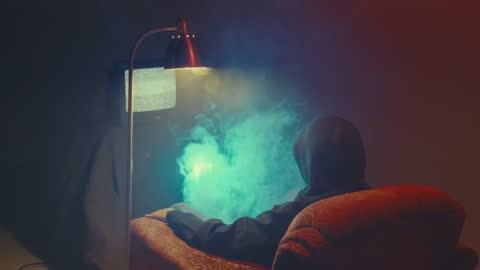 RUINAS BTS, frame 06: a seated figure in an armchair in teal smoke under a lamp