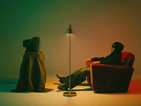 RUINAS, frame 06: a hooded figure in an armchair beside a floor lamp in a green room