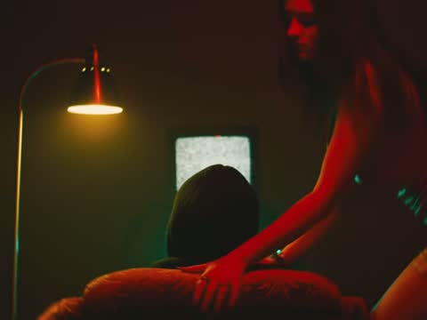 RUINAS, frame 14: a lamp, a TV and a woman leaning over a couch in a dark red room