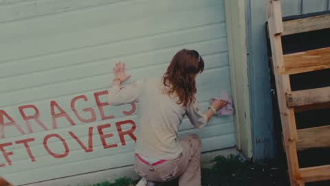 SPCA, frame 02: a woman painting red lettering on a white garage wall
