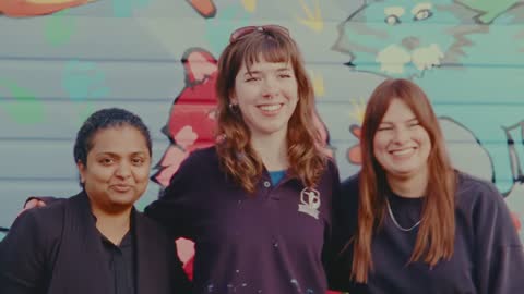 SPCA, frame 13: three women smiling in front of the mural